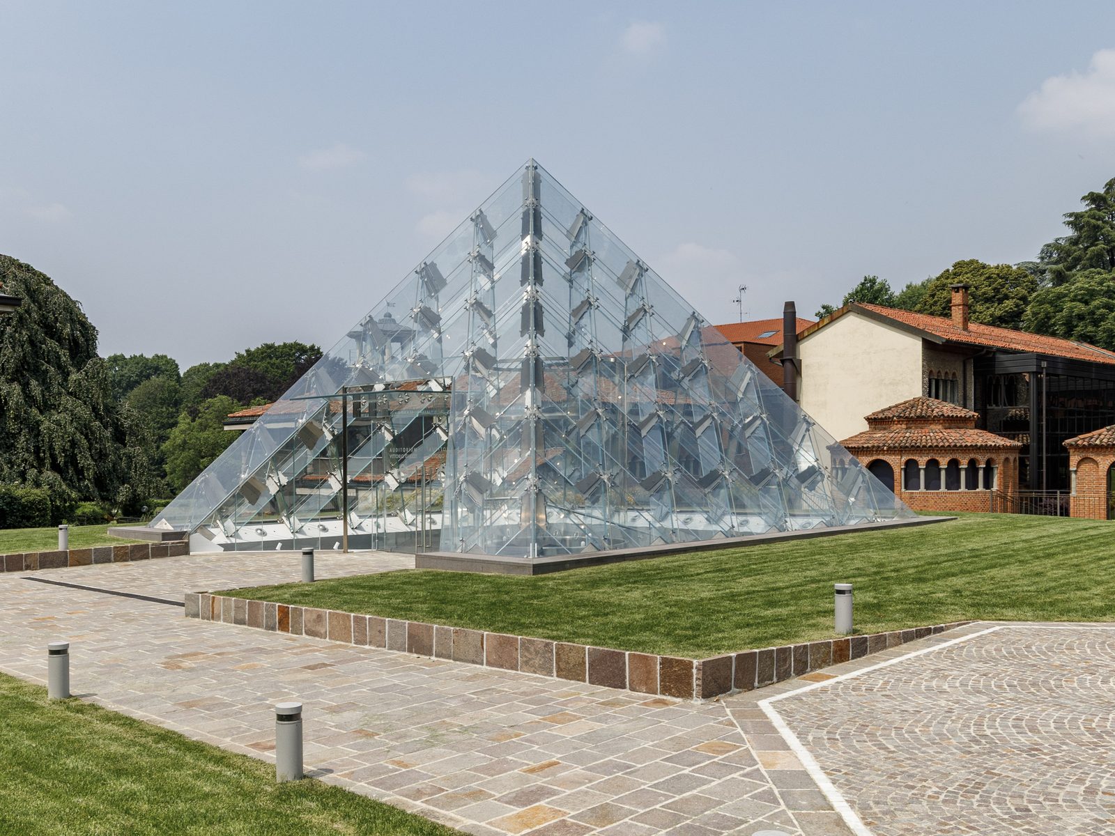 Vista ampia della Glass Pyramid a Carate Brianza nel parco, con edifici storici in mattoni sullo sfondo